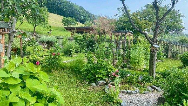 Ein gepflegter Garten mit Blumen, Bäumen und einem Kiesweg, umgeben von einem Holzzaun und einer grünen Landschaft im Hintergrund.