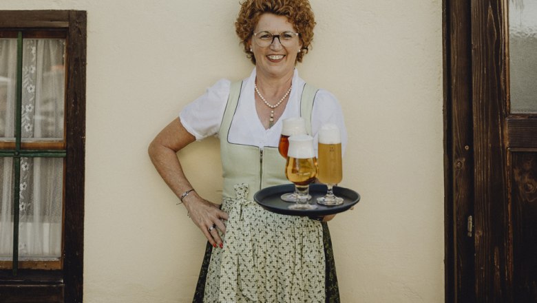 Frau in traditioneller Kleidung mit Tablett voller Biergläser vor einer Wand.