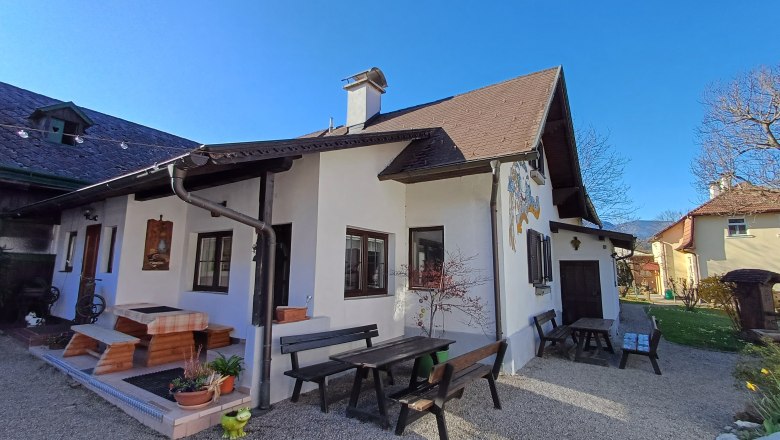 Ein traditionelles Haus mit Holzbänken und Tischen im Garten, umgeben von blühenden Pflanzen und Bäumen unter einem klaren blauen Himmel.