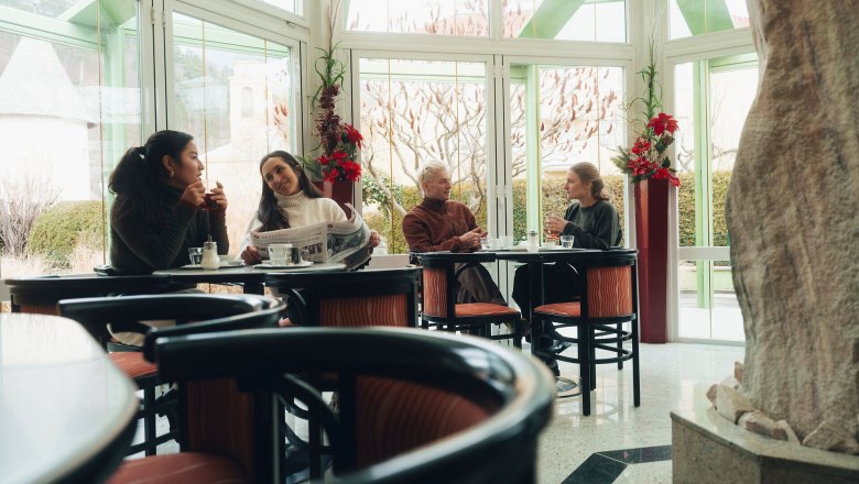 In der gemütlichen Atmosphäre der Konditorei Nöbauer genießen die Gäste köstliche Leckereien und anregende Gespräche. Die großen Fenster bieten einen herrlichen Blick auf die winterliche Landschaft der Wiener Alpen, während die warmen Farben des Interieurs eine einladende Stimmung schaffen.
