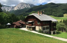 Ein traditionelles Holzhaus in einer bergigen Landschaft mit gr&uuml;nen Wiesen und bewaldeten H&uuml;geln im Hintergrund.
