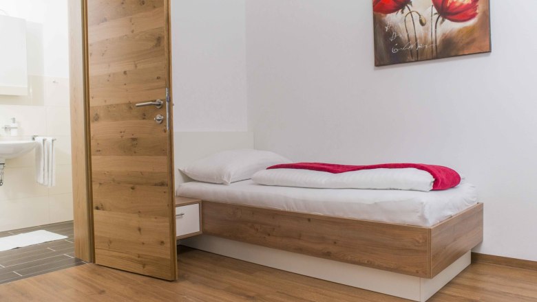 Ein einfach eingerichtetes Zimmer mit einem Einzelbett, Holzboden und einem Bild mit roten Blumen an der Wand.