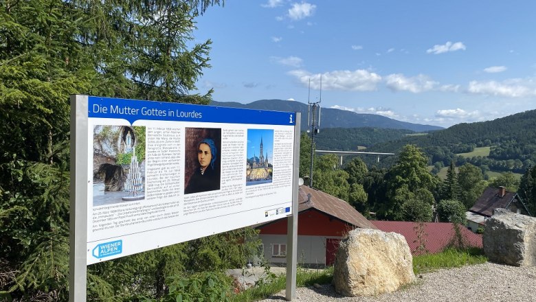 Informationsschild über die Mutter Gottes in Lourdes vor einer Landschaft mit Hügeln und Bäumen.
