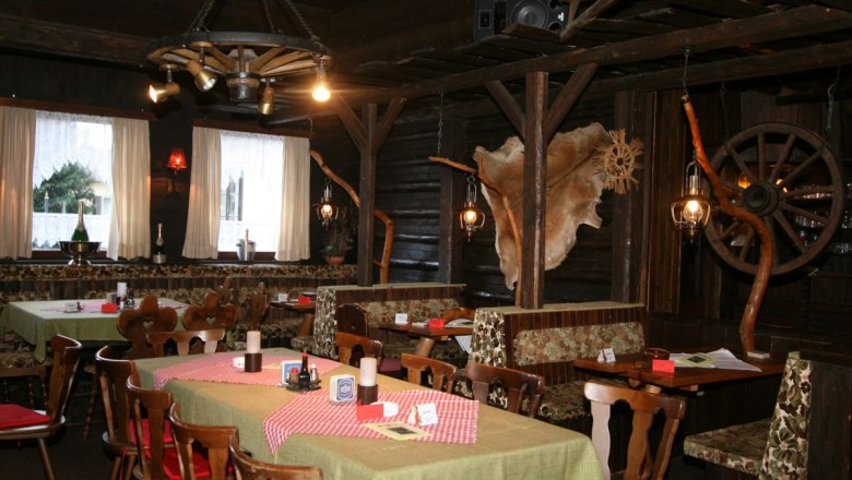 Rustikales Restaurant mit Holzm&ouml;beln, karierten Tischdecken und dekorativen Elementen wie Wagenr&auml;dern und Tierfell an den W&auml;nden.