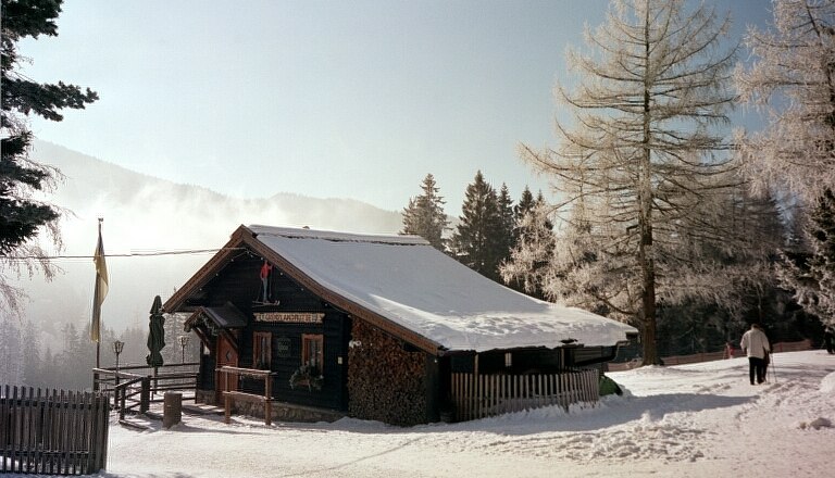 Eine verschneite Bergh&uuml;tte in winterlicher Landschaft mit B&auml;umen und einem Spazierg&auml;nger.