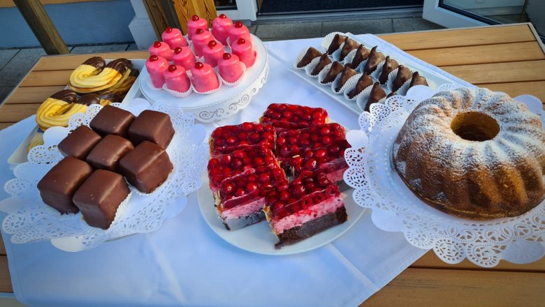 Verschiedene hausgemachte Mehlspeisen auf einem Tisch, darunter Gugelhupf, Schokoladenwürfel und Kirschkuchen.