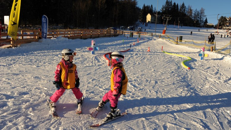 Zwei Kinder in Skiausrüstung im Schnee, umgeben von bunten Hindernissen.