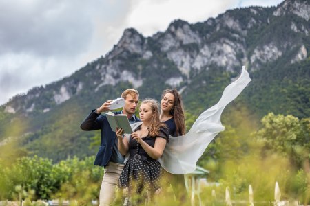 drei junge Personen, die ein Buch und eine Maske in der Hand haben und auf einer Wieser in bergiger Landschaft stehen