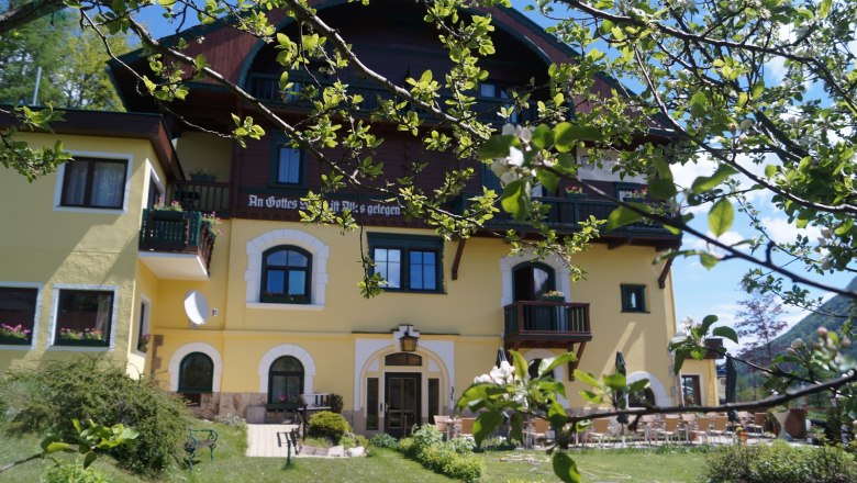 Gartenansicht des Hotel Belvedere mit gelber Fassade und blühenden Bäumen im Vordergrund.