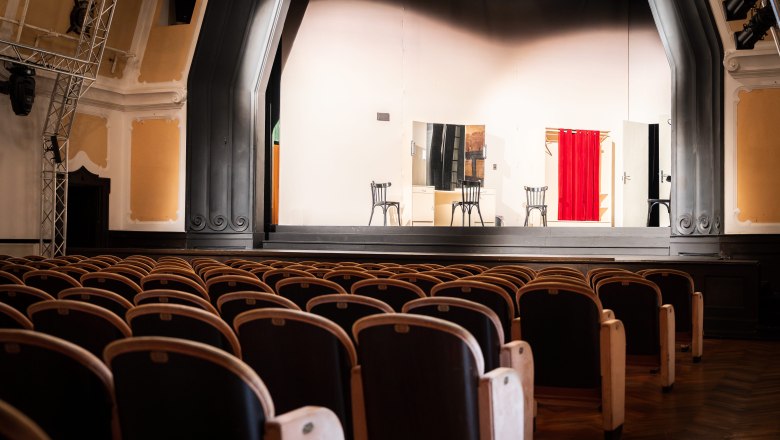 Leerer Theatersaal mit Bühne, auf der zwei Stühle und ein roter Vorhang zu sehen sind.