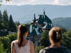 Zwei Menschen, mit dem R&uuml;cken zur Kamera, blicken auf eine malerische Berglandschaft. Im Vordergrund sieht man das S&uuml;dbahnhotel, eingebettet in &uuml;ppiges Gr&uuml;n. Im Hintergrund erheben sich Berge unter einem klaren blauen Himmel.