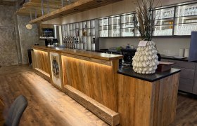 Holztheke in einem Restaurant mit dekorativer Vase und Gl&auml;serregal im Hintergrund.