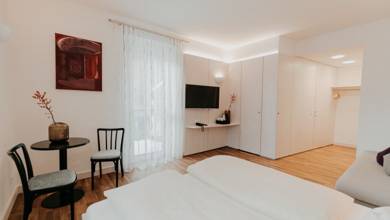 Modernes, helles Hotelzimmer mit Doppelbett, TV, Tisch und Stühlen, Schrank und dekorativem Bild an der Wand.