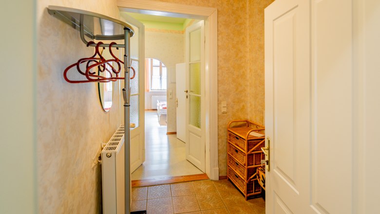 Ein Vorraum mit Kleiderhaken, einem Heizk&ouml;rper und einem Korbm&ouml;bel. Eine T&uuml;r f&uuml;hrt in einen hellen Raum mit Fenstern.