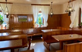 Gemütliche Gaststube mit Holzmöbeln, weißen Vorhängen und Pflanzen auf der Fensterbank.