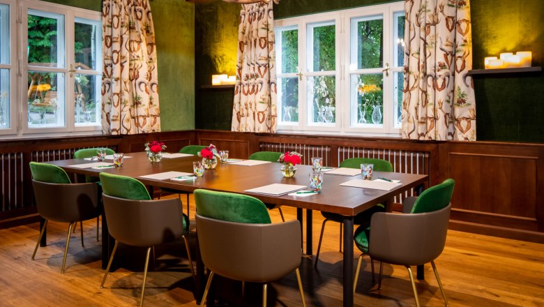 Ein Jagdzimmer mit Holztisch, gr&uuml;nen St&uuml;hlen und Geweihleuchter.