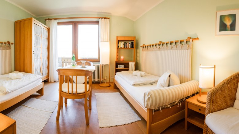 Gemütliches Zweibettzimmer mit Holzmöbeln, zwei Betten, Tisch, Stühlen und einem Fenster mit Vorhängen.