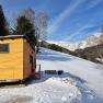 Kleines Holzhaus auf schneebedecktem H&uuml;gel mit Berglandschaft im Hintergrund.