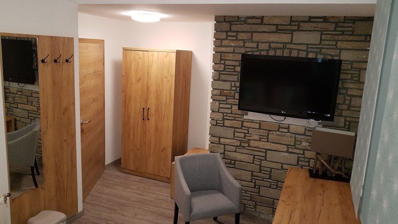 Hotelzimmer mit Holzmöbeln, grauem Sessel und Flachbildfernseher an einer Steinwand.