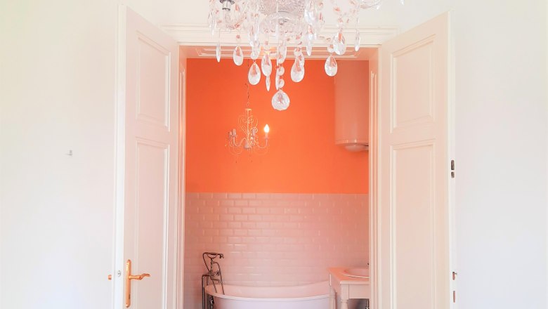 Blick in ein Badezimmer mit freistehender Badewanne, orangefarbener Wand und Kronleuchter.