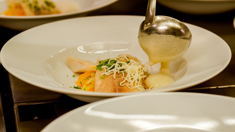 Ein Teller mit Fisch, Gem&uuml;se und einer hellen So&szlig;e, die mit einer Kelle serviert wird.