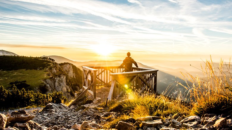 Person steht auf einer Holzplattform und blickt in den Sonnenuntergang über einer Berglandschaft.