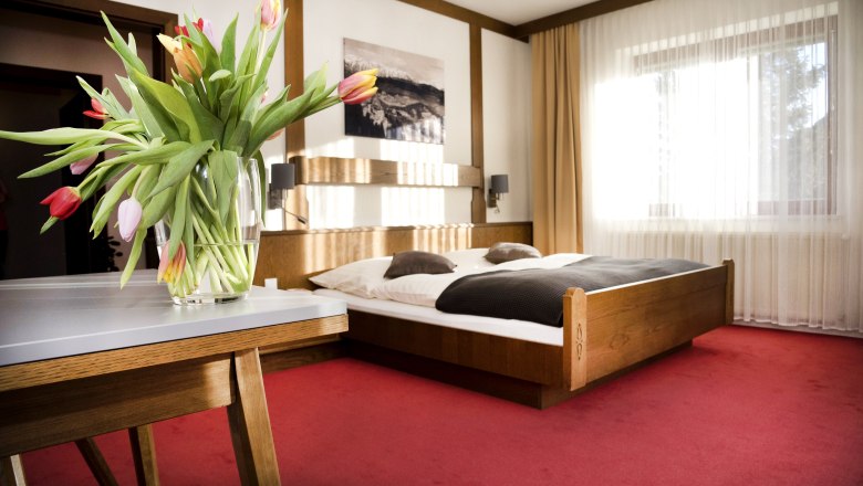 Ein gem&uuml;tliches Doppelzimmer mit einem Holzbett, roten Teppichboden und einem Tisch mit einem Blumenstrau&szlig; aus Tulpen.