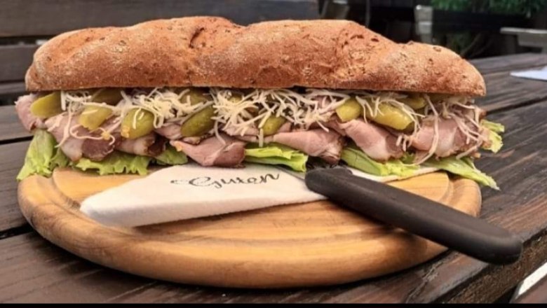 Ein großes Sandwich mit Fleisch, Salat, Gurken und Käse auf einem Holzbrett, daneben ein Messer mit Serviette.