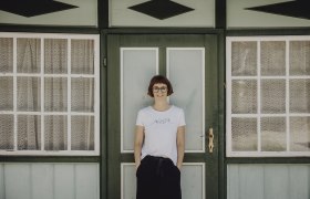 Eine Frau steht vor einer grünen Tür mit Fenstern und Spitzenvorhängen.