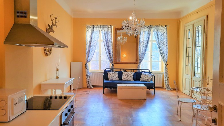 Wohnzimmer mit K&uuml;che, Esstisch und Sofa, gelbe W&auml;nde, Kronleuchter, Fenster mit Vorh&auml;ngen.