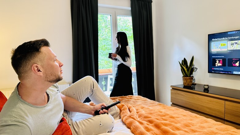 Ein Mann sitzt auf einem Bett und schaut fern, während eine Frau am Fenster steht und Kaffee trinkt.