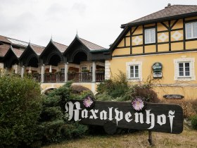 Traditionelles Gasthaus mit gelber Fassade und Holzveranden, davor ein Schild mit der Aufschrift &bdquo;Raxalpenhof&ldquo;.
