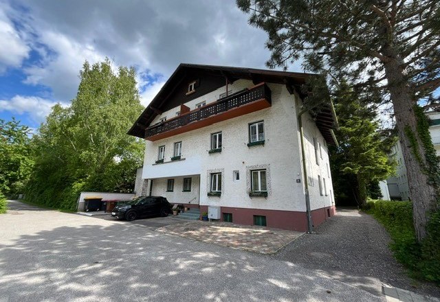 Zweistöckiges Haus mit weißer Fassade und dunklem Dach, umgeben von Bäumen und einem gepflasterten Hof.