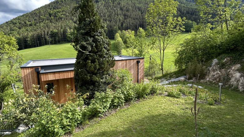 Holzhaus mit Garten vor bewaldetem Hügel und blauem Himmel.