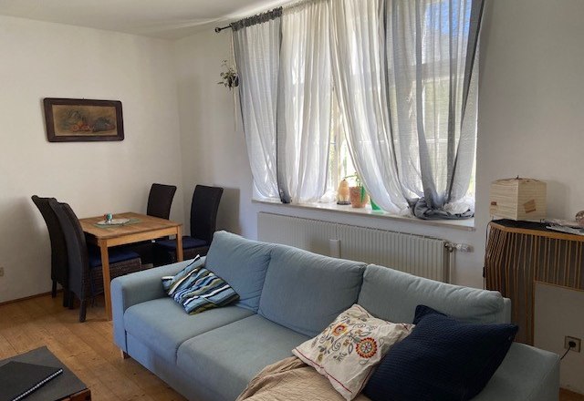 Ein Wohnzimmer mit einem blauen Sofa, einem Holztisch mit Stühlen und einem Fenster mit weißen Vorhängen.