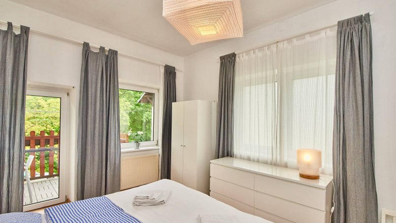 Helles Schlafzimmer mit Doppelbett, blauen Streifenbettwäsche, weißen Möbeln und grauen Vorhängen. Fenster mit Blick auf einen Balkon.