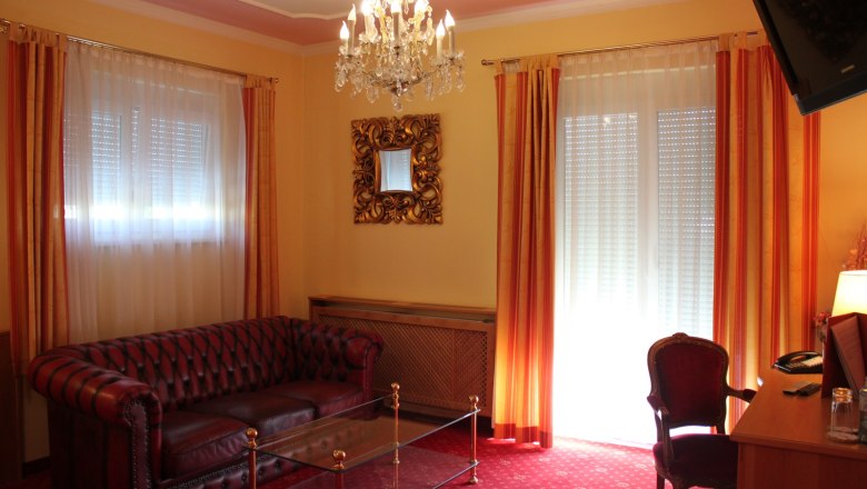 Elegantes Wohnzimmer mit Ledersofa, Glastisch, Kronleuchter und roten Vorhängen.