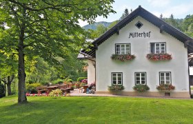 Ein traditionelles Bauernhaus mit dem Namen Mitterhof, umgeben von gr&uuml;ner Landschaft und Blumenbeeten.