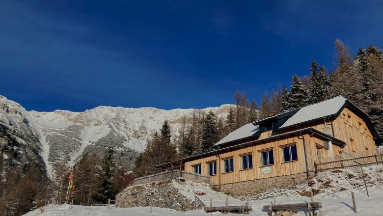 Ein Holzhaus im Schnee vor einer Bergkulisse unter klarem, blauem Himmel.