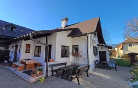 Ein traditionelles Haus mit Holzb&auml;nken und Tischen im Garten, umgeben von bl&uuml;henden Pflanzen und B&auml;umen unter einem klaren blauen Himmel.