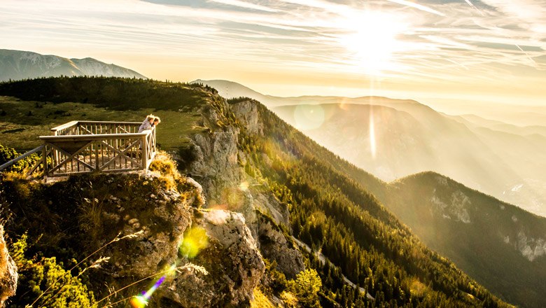 Aussichtsplattform auf einem Berg mit Blick auf bewaldete Hügel und Sonnenuntergang.