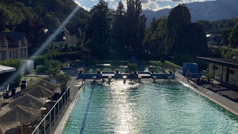 Ein Freibad mit klarem Wasser, umgeben von B&auml;umen und Bergen im Hintergrund.