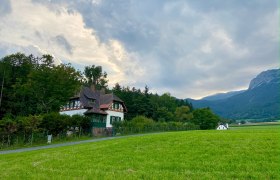 Ein Haus im traditionellen Stil steht vor einem bewaldeten Hügel, umgeben von grünen Wiesen und Bergen im Hintergrund.