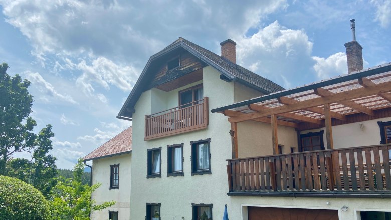 Ein zweistöckiges Haus mit Balkon und Terrasse, umgeben von Garten und Bäumen, unter einem bewölkten Himmel.