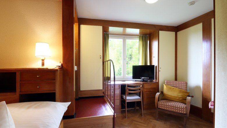 Ein gemütliches Hotelzimmer mit Holzmöbeln, einem Fernseher, einem Sessel und einem Fenster mit Blick ins Grüne.