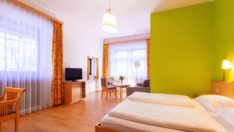 Helles Hotelzimmer mit Doppelbett, grünem Akzent an der Wand, Fernseher, Sitzbereich und großen Fenstern mit Vorhängen.