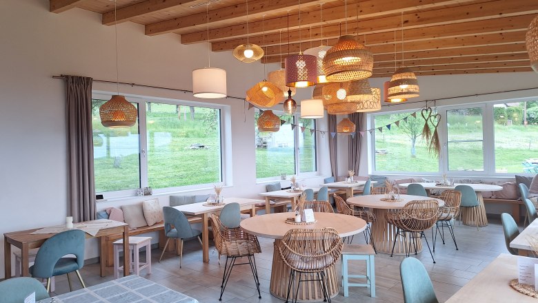Gemütlicher Gastraum mit Holzmöbeln und Hängelampen, große Fenster mit Blick ins Grüne.