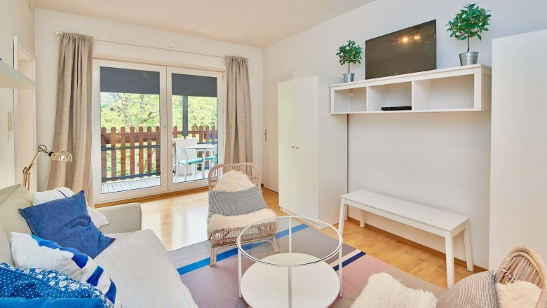 Modernes Wohnzimmer mit Sofa, Sessel, Glastisch und TV. Balkon mit Holzzaun im Hintergrund.