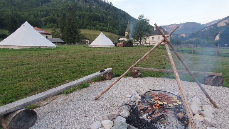Lagerfeuer mit Grill &uuml;ber Feuerstelle, Zelte im Hintergrund, umgeben von Bergen und Wiesen.