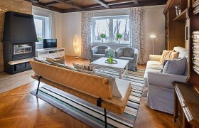 Gemütliches Wohnzimmer mit Kamin, Sofa, Sessel und Fernseher. Holzboden und Decke, gestreifter Teppich, große Fenster mit Vorhängen.